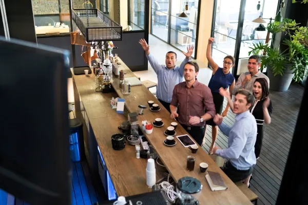 A group of six diverse office workers in a modern break room stand behind a counter, cheering and raising their arms in excitement while watching something off-camera.